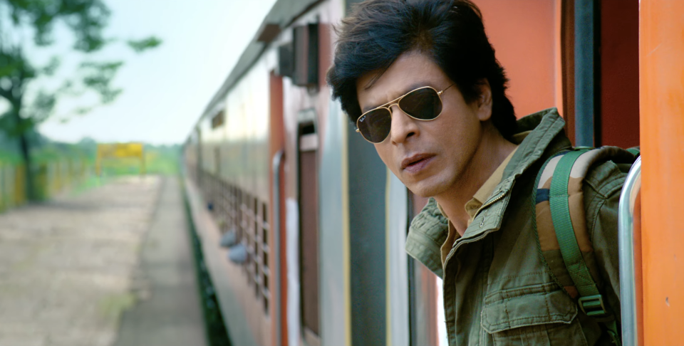 A man in aviator sunglasses leaning out of an orange moving rain; Shah Rukh Khan in "Dunki"