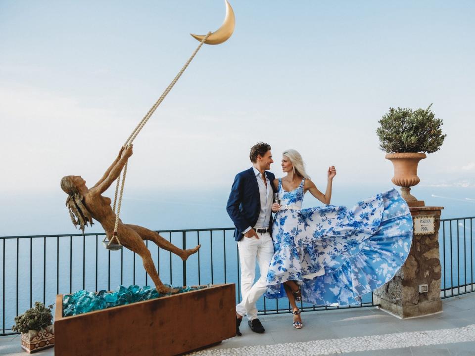 A bride and groom look at each other in front of a balcony overlooking the ocean as the woman's dress flows in the wind.