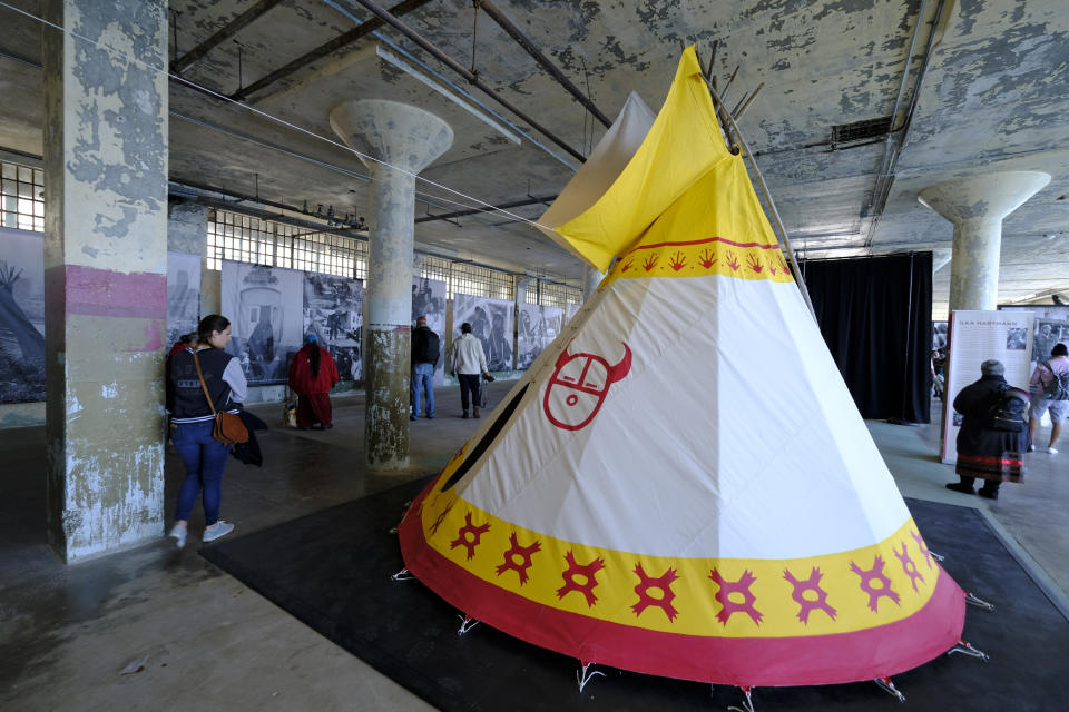 People make their way past a teepee and large photographs that are part of an exhibit for the 50th anniversary of the Native American occupation on Alcatraz Island Wednesday, Nov. 20, 2019, in San Francisco. About 150 people gathered at Alcatraz to mark the 50th anniversary of a takeover of the island by Native American activists. Original occupiers, friends, family and others assembled Wednesday morning for a program that included prayer, songs and speakers. They then headed to the dock to begin restoring messages painted by occupiers on a former barracks building. (AP Photo/Eric Risberg)