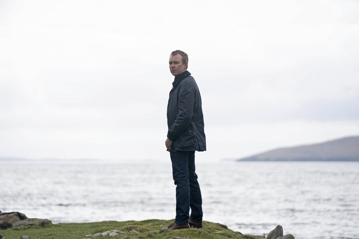  Shetland's DC Sandy Wilson (Steven Robertson) stands on a cliff edge with the ocean behind him. His body is turned away from the camera but he is looking back over his shoulder at us. 