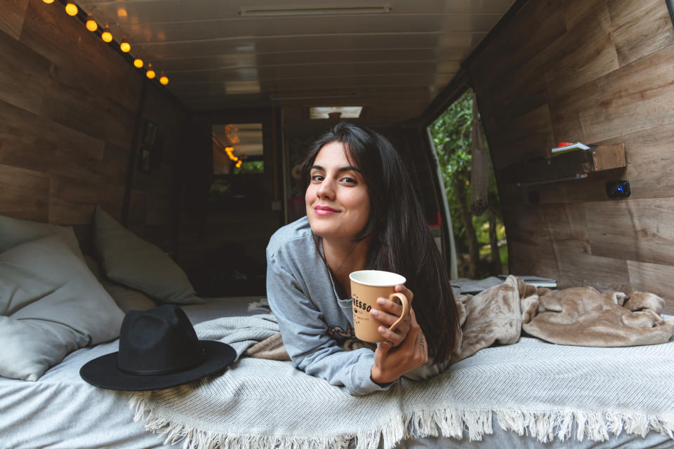 Woman lying on a bed in a cozy, wooden-paneled van, smiling and holding a mug. A hat is beside her on the bed