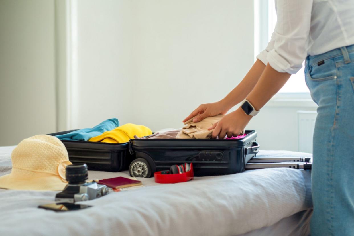 Mid-section of woman preparing small carry-on bag on bed, focus on items to be put in bag, bed, white walls, and a window in the background