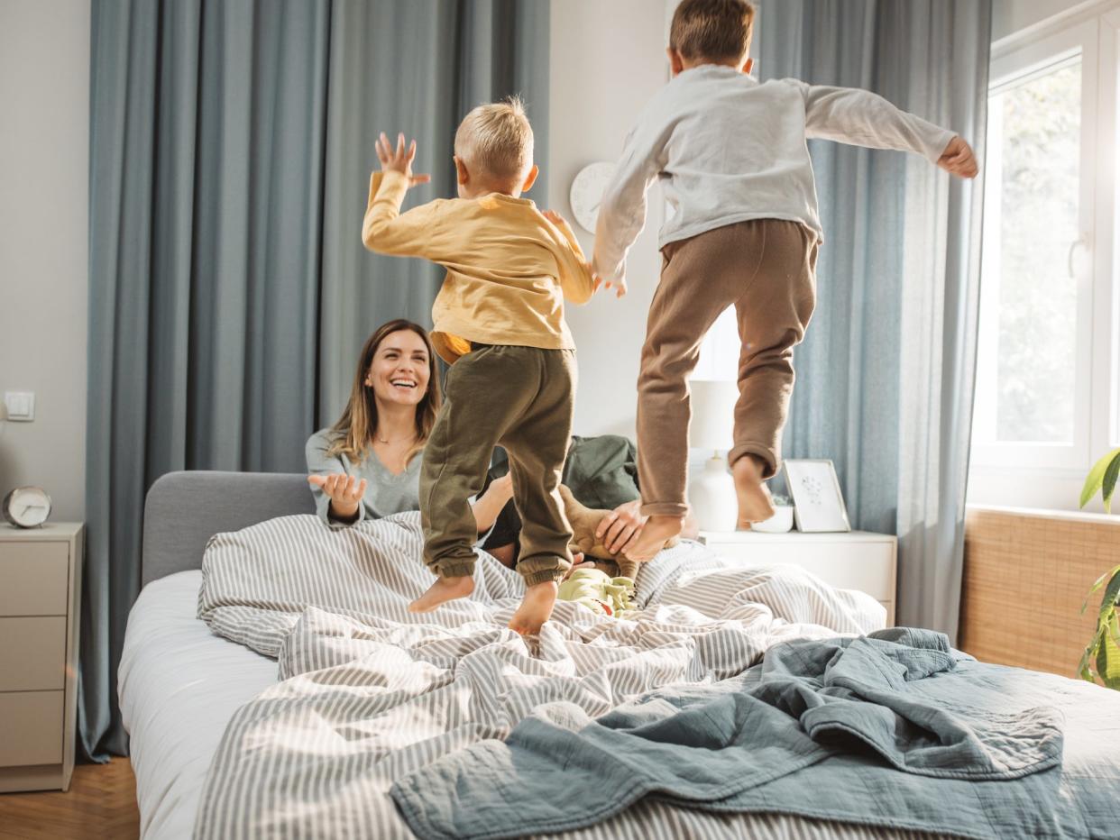 Family of four in bed in the morning. Two young boys are jumping on the bed and the mother is smiling at them.