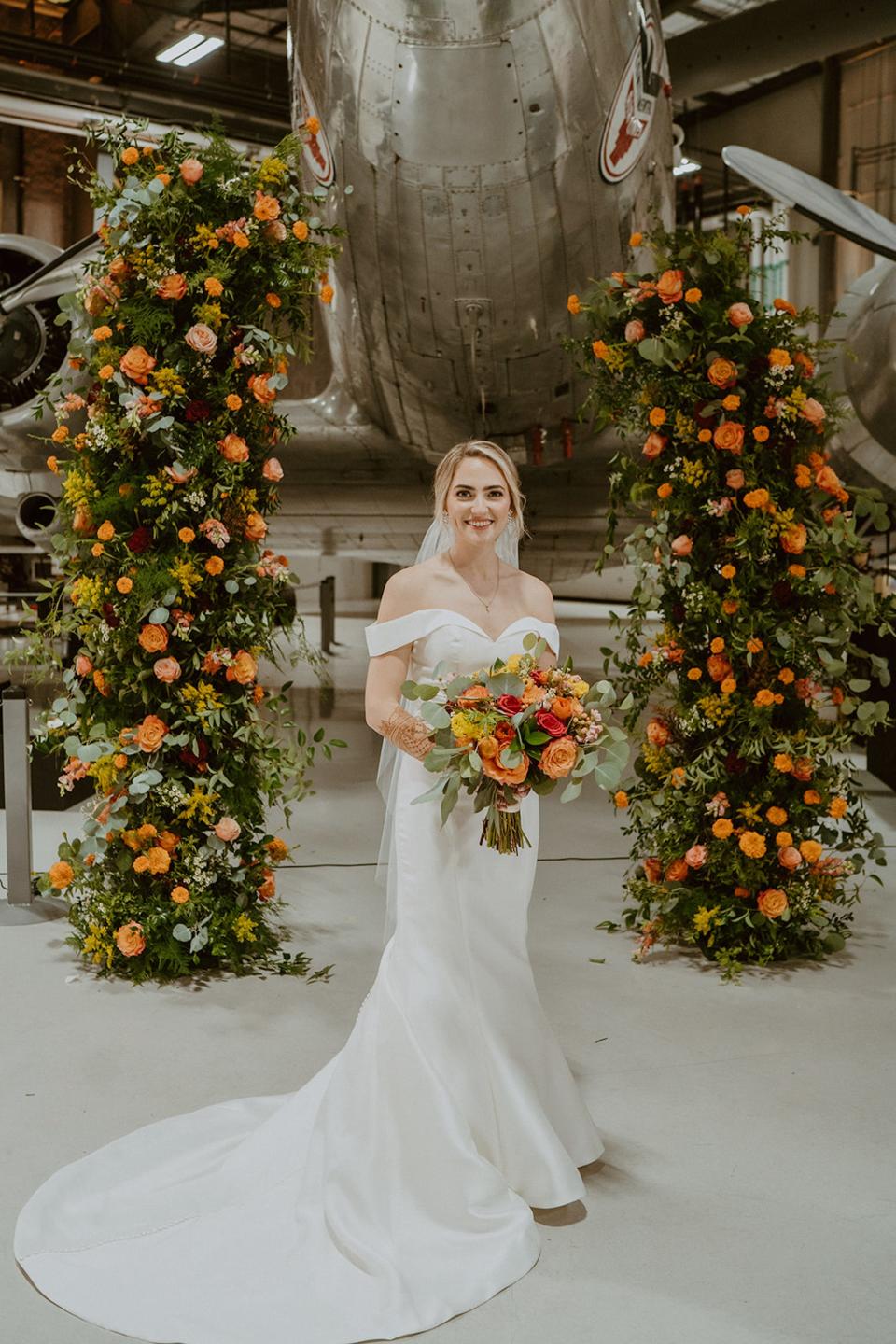 A bride in a white wedding dress stands in front of a floral archway and a plane.