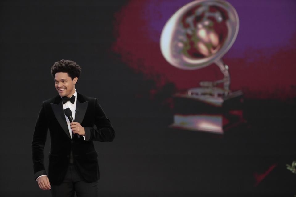 A man in a tuxedo holding a microphone onstage. A Grammy trophy is projected behind him.