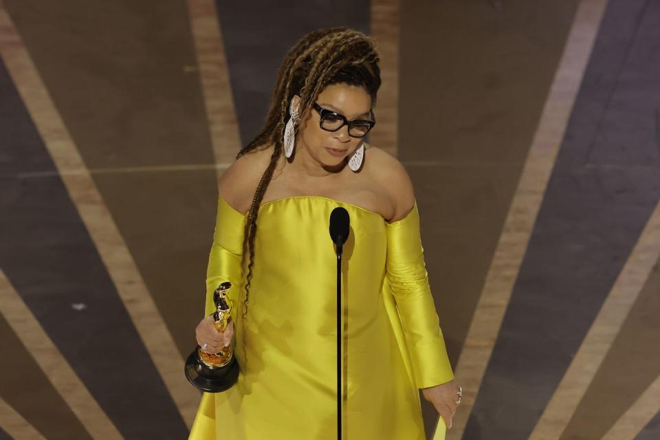 ruth e carter, wearing a yellow dress, white earrings, and black glasses, holds an academy award and speaks into a microphone on a stage