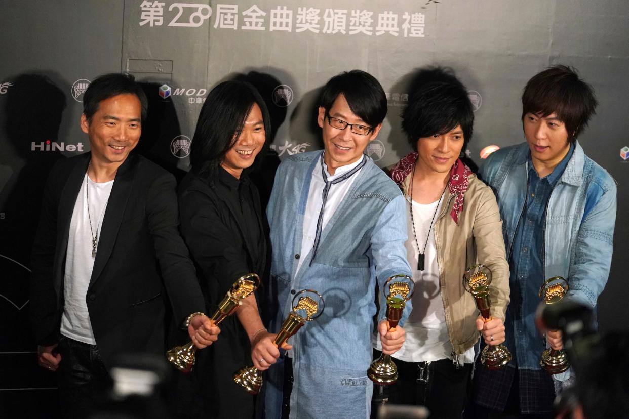 Five men who are a part of the band Mayday hold gold trophies with their arms outstretched as they smile to the camera