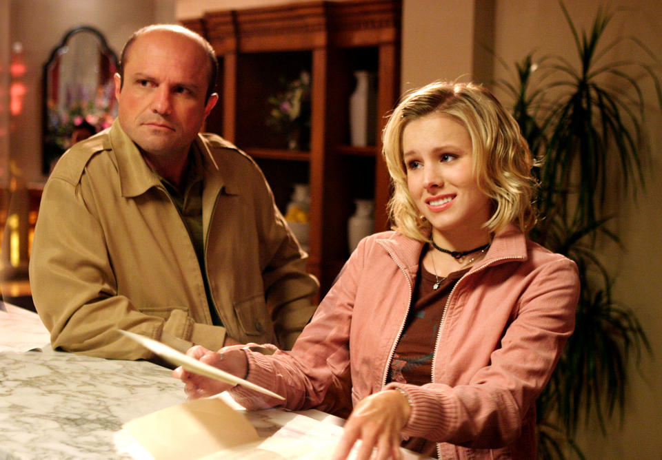Enrico Colantoni and Kristen Bell in a scene, standing at a counter. Kristen holds a paper and smiles, while Enrico looks concerned