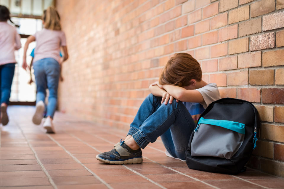 Little boy sitting alone on floor after suffering an act of bullying while children run in the background. Sad young schoolboy sitting on corridor with hands on knees and head between his legs.