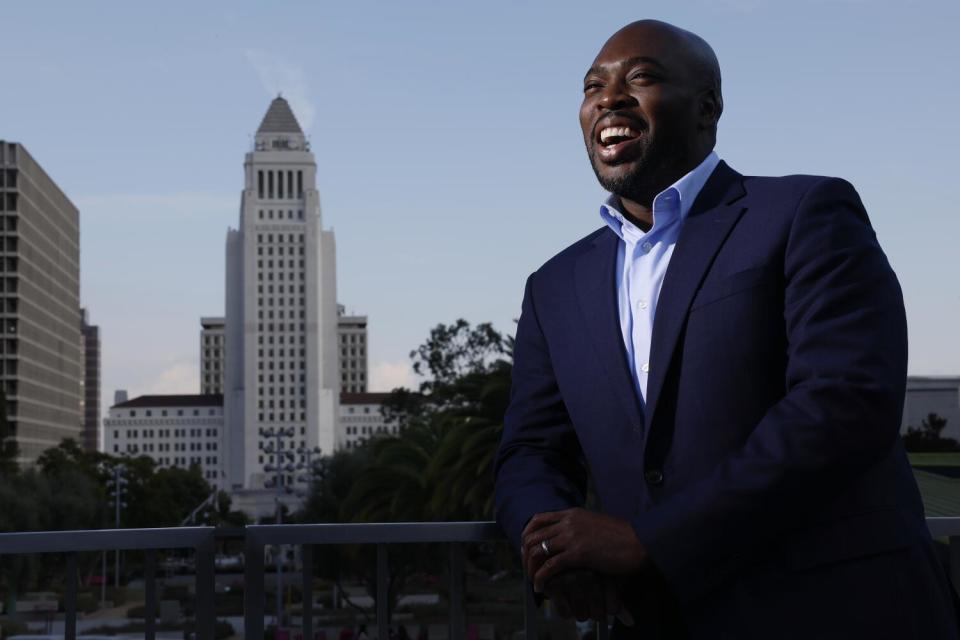 A man pictured from the waist up, standing, with Los Angeles City Hall and trees in the background