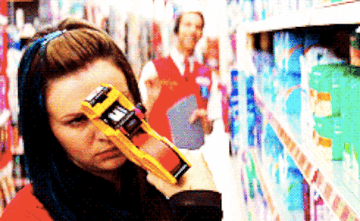 A woman scans products in a store aisle with a barcode scanner, while a man in a red vest stands in the background holding a clipboard