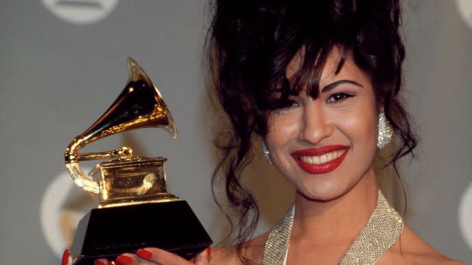 selena quintanilla smiles at the camera and holds her grammy award, she wears a sparkling gown and large sparkling earrings