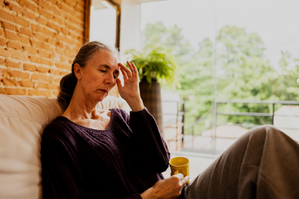 A woman with graying hair, seated on a couch, looks pensive while holding a yellow mug in one hand and touching her forehead with the other