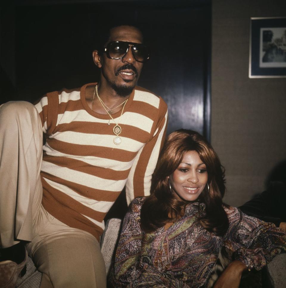 ike turner and tina turner sit on a couch next to each other, ike wears an orange and white striped shirt with khakis, gold necklaces and sunglasses, tina wears a paisley top and hoop earrings