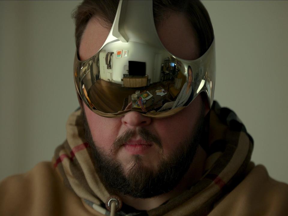 jack rooney in 3 body problem, wearing a metallic headset over his eyes and ears that reflects the world around him. he has a neutral expression on his face underneath the head piece