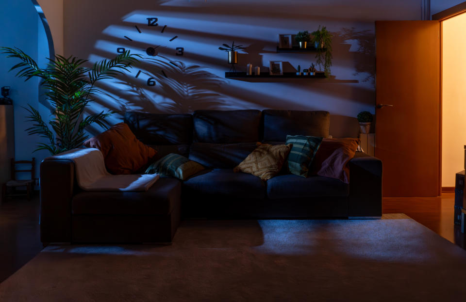 A cozy living room with a large sofa adorned with various pillows under a wall clock and shelves holding plants and decor, illuminated by soft lighting