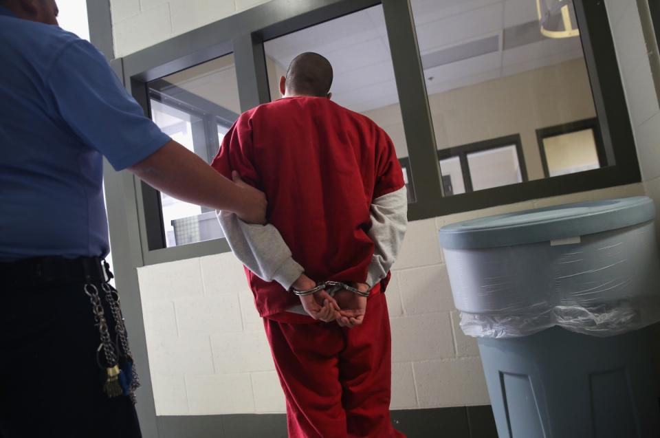 A man in red jumpsuit and handcuffs is escorted by a guard