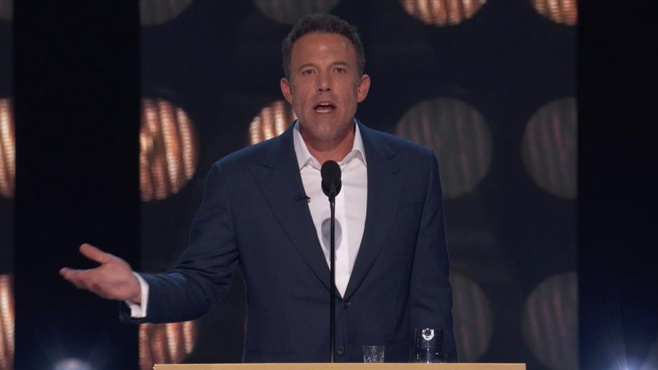 Ben Affleck in a navy suit stands behind the podium during the Tom Brady roast looking angry