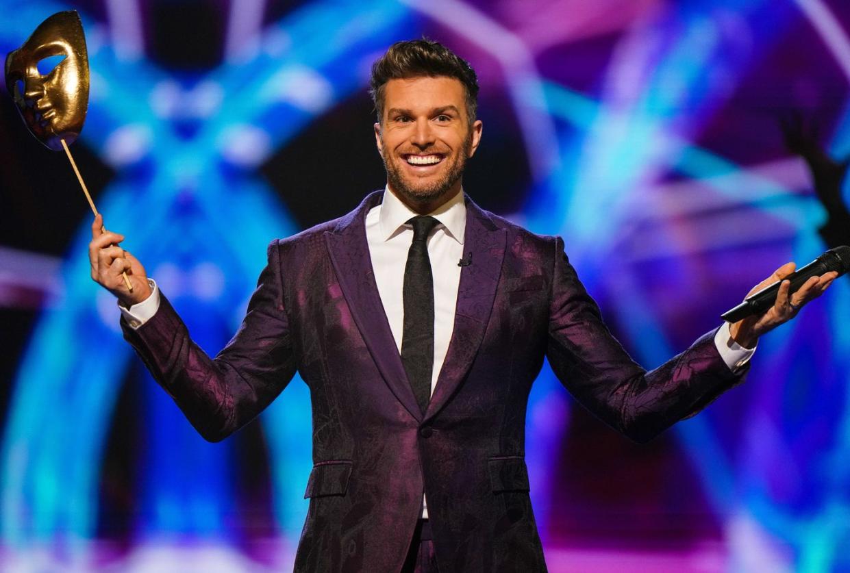 the masked dancer host joel dommett on stage, holding a gold theatre style face mask on a stick in his right hand and his microphone in his left hand