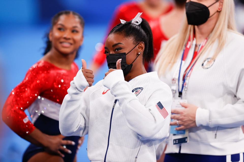 Shows Biles, wearing a mask and a white warm-up suit, giving the thumbs-up.