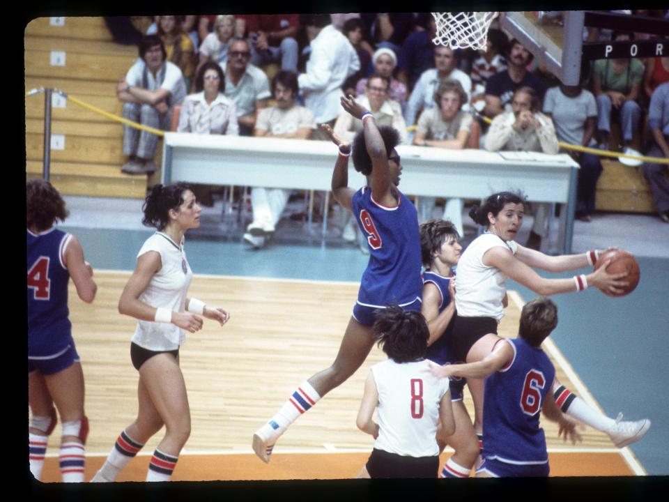 A group of women play basketball with a crowd in the background at the 1976 Olympics.