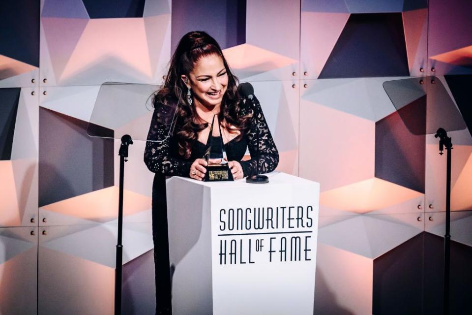 gloria estfan smiles as she stands at a podium and touches a crystal award resting on the podium, she wears a black long sleeve dress