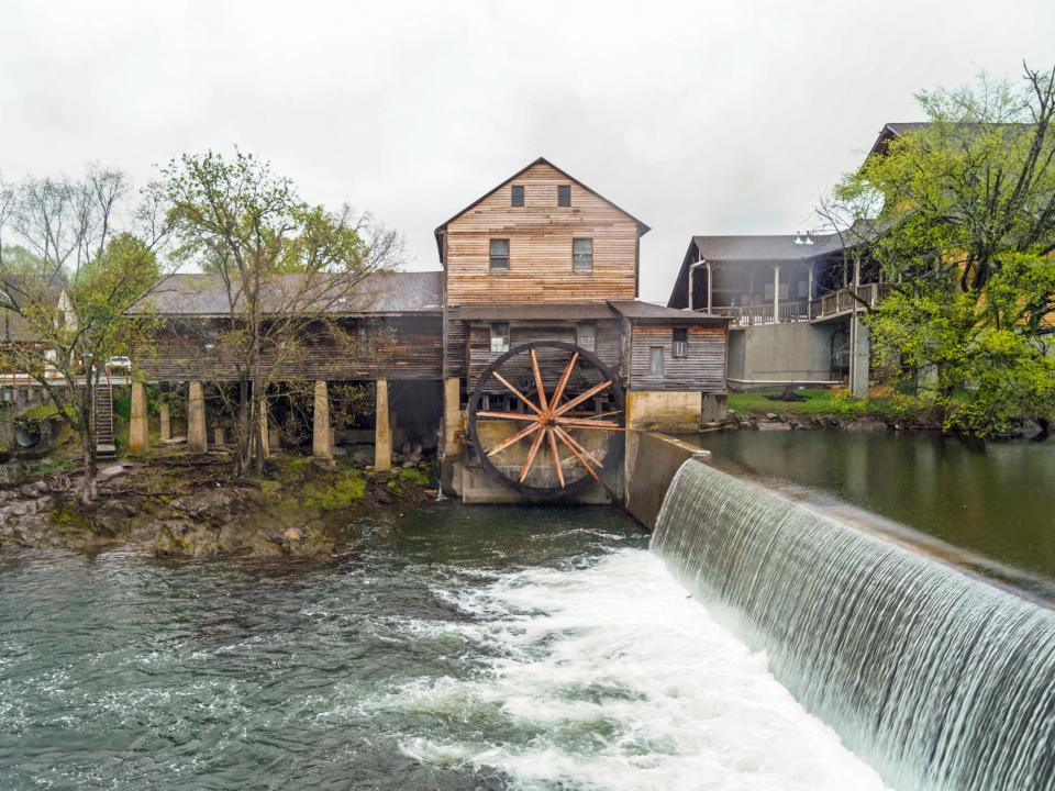 A dam waterfall on the left and a wooden mill building behind it with cloudy skies