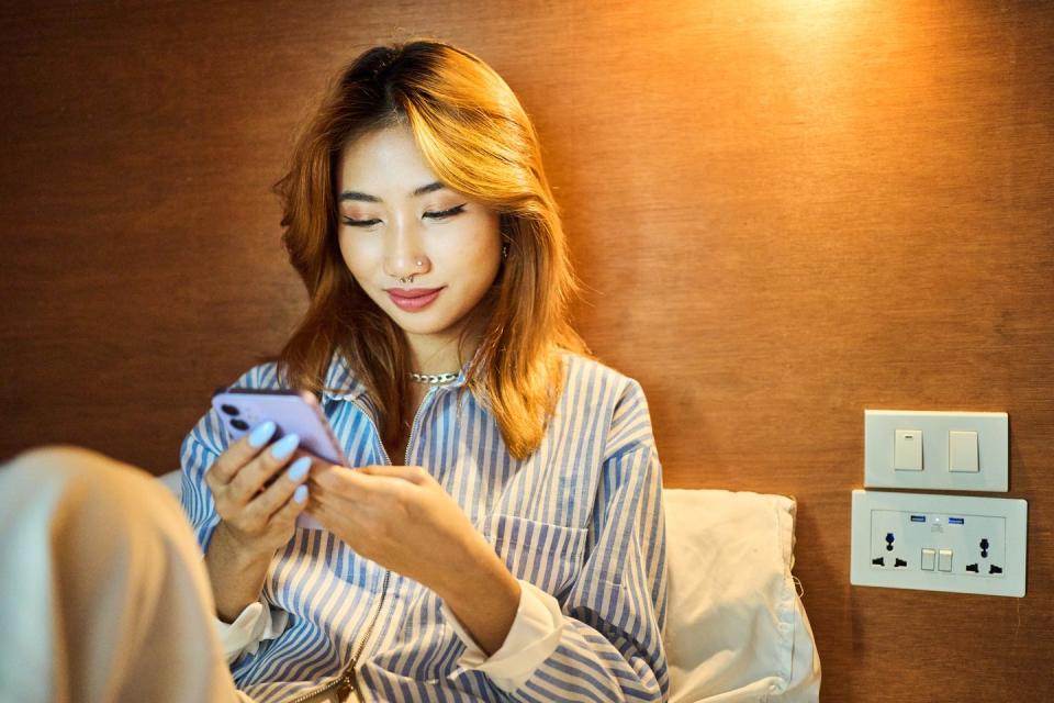 smiling young woman checking her smart phone while relaxing in her bunk in a hostel dorm during a vacation