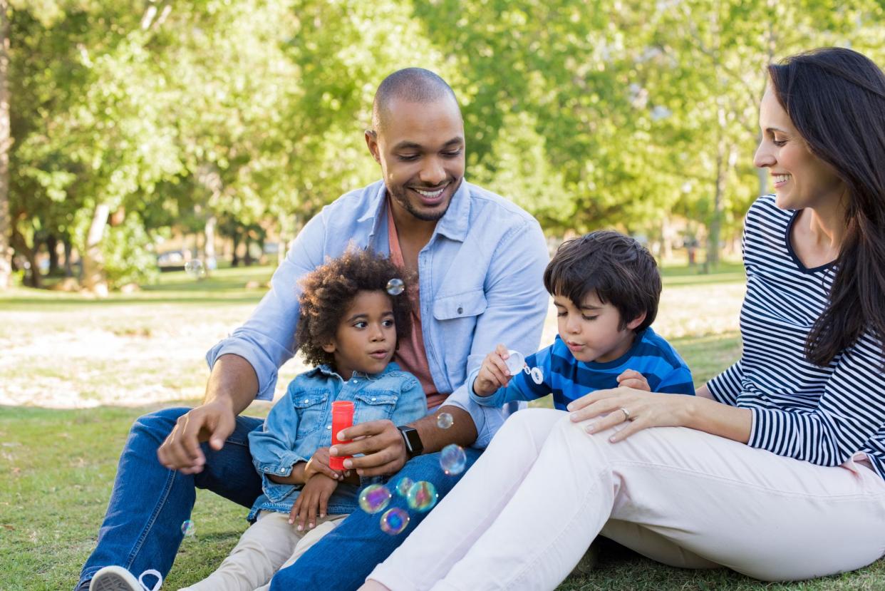 Happy family smiling and having fun outdoors. Young latin boy with brother and parents playing with soap bubbles at park. Multiethnic parents watching children enjoying making soap bubbles.