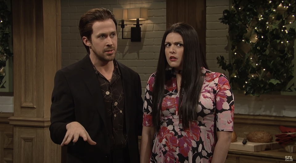 Ryan Gosling in a patterned shirt and jacket stands next to Cecily Strong, who is in a floral dress, during an SNL skit in a comedy restaurant setting