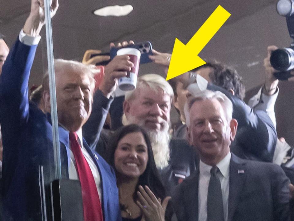 Donald Trump and Melania Trump smile and wave with a crowd of people, some taking photos, behind a glass barrier at an event