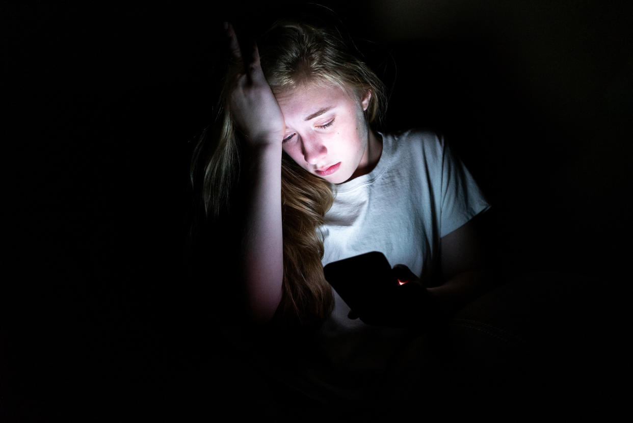 A girl looking distressed sits in the dark, with her smartphone's screen lighting her face. (Getty Images)