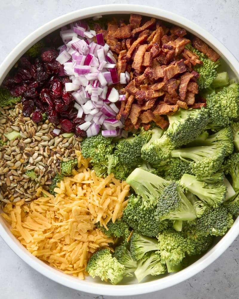 overhead shot of all the ingredients of the broccoli salad organized in a circle around a large white bowl.