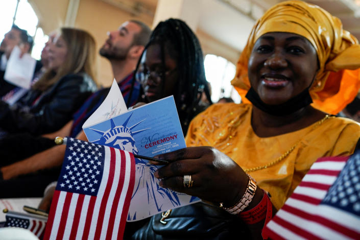 A woman in an orange headdress, seated with other prospective citizens, holds a program for the ceremony and a flag. 