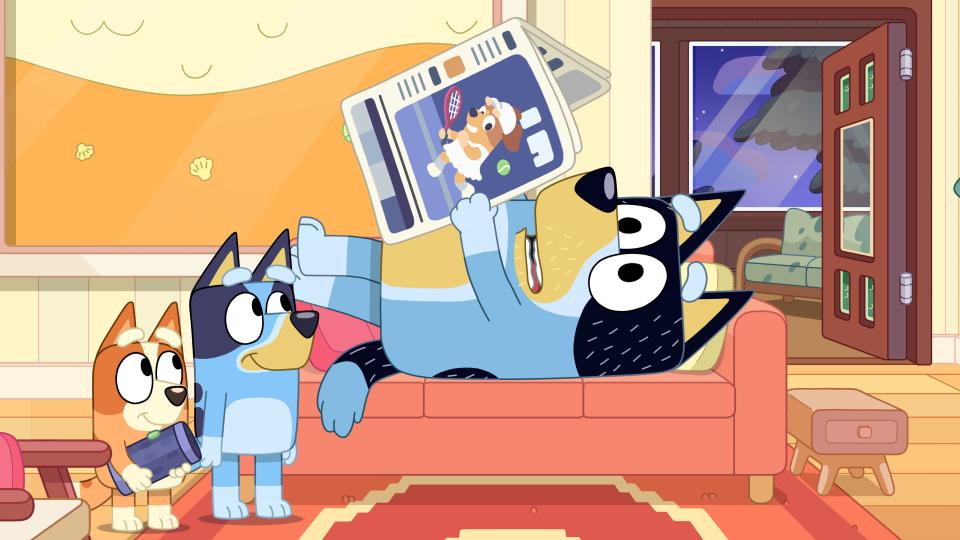 Bandit "Dad" reading the newspaper on the couch while Bluey and Bingo wait for him to play music on his phone.