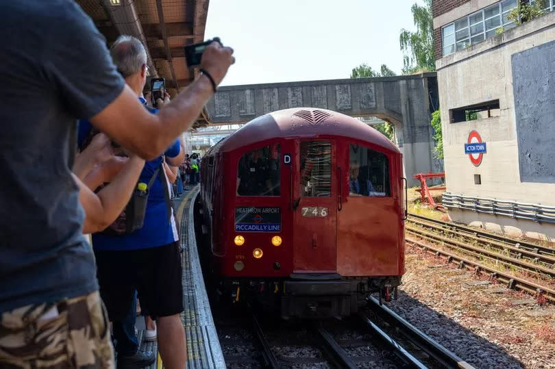 A 1938 Art Deco-style London underground train arrives at Acton Town station as people take photos