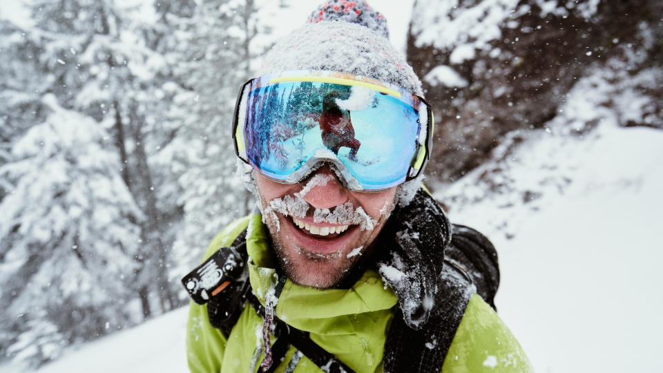 A skier wearing goggles is smiling and snow covered on a powder day