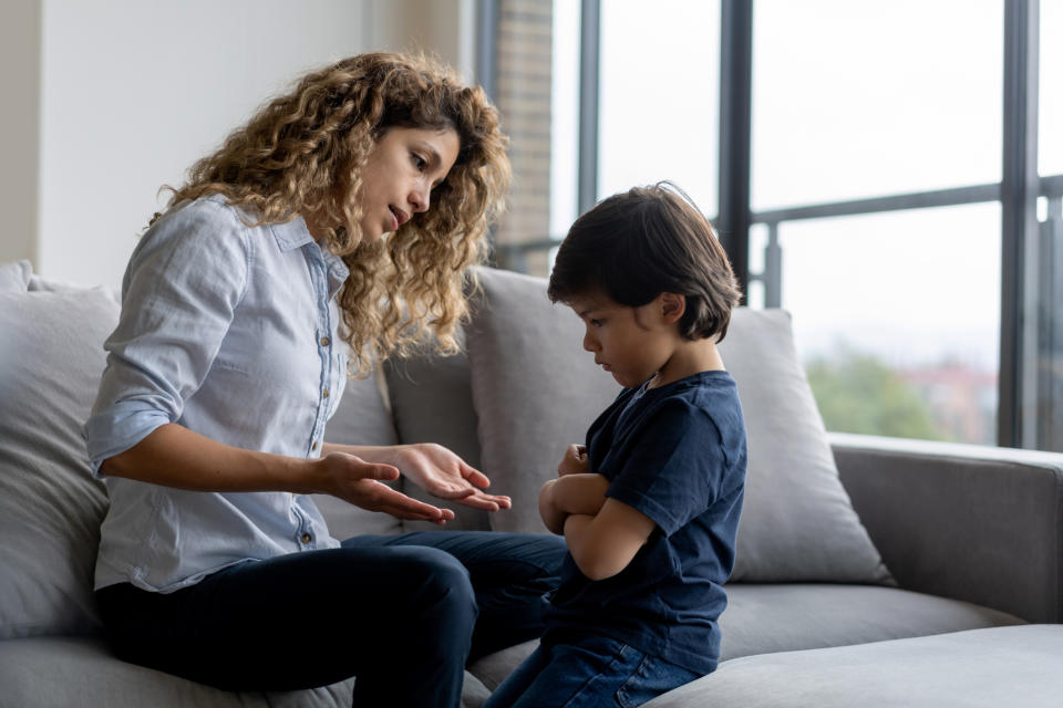 An adult with curly hair talks to a young child who looks down, arms crossed, in a living room