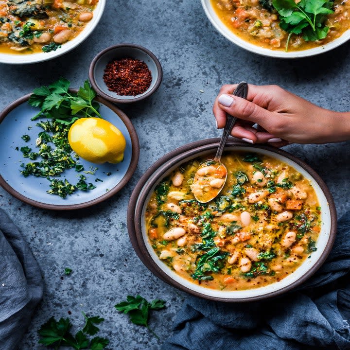 Three bowls of white bean and kale soup with fresh herbs, lemon juice, and red pepper flakes on top.