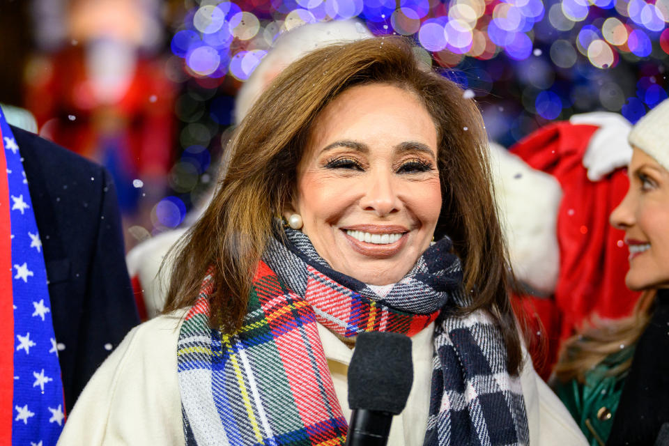 Jeanine Pirro smiles, wearing a checkered scarf and holding a microphone at a festive event with blurry Christmas lights in the background