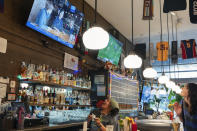 Bartender M.J. Jones, left, and fellow employee Allison Clarke, right, react as an interview with The Sports Bra founder and CEO Jenny Nguyen, center, airs on a screen during their shift at the sports bar on Wednesday, April 24, 2024, in Portland, Ore. A painting of U.S. soccer legend Abby Wambach is mounted above the chalkboard beer menu. (AP Photo/Jenny Kane)