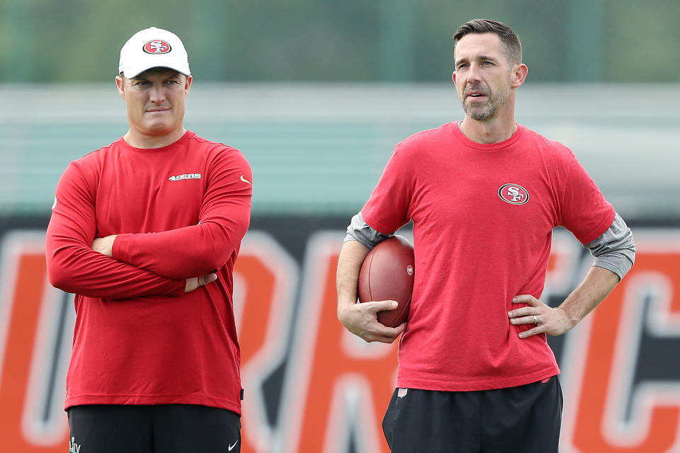 John Lynch, left, and Kyle Shanahan stand next to each other in red 49ers shirts as Shanahan holds a football.