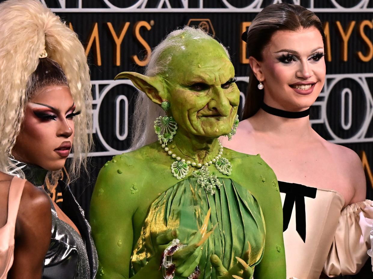 drag race queens on the emmys red carpet, with a person dressed as a goblin in a green dress, warts on her skin, her hair stringly, her face withered