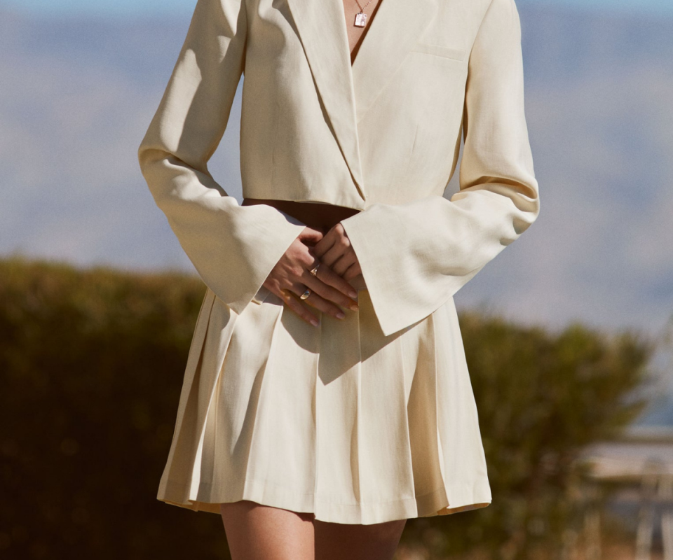 A young woman wears a cream pleated mini skirt with a matching blazer with trees and blue skies in the background.