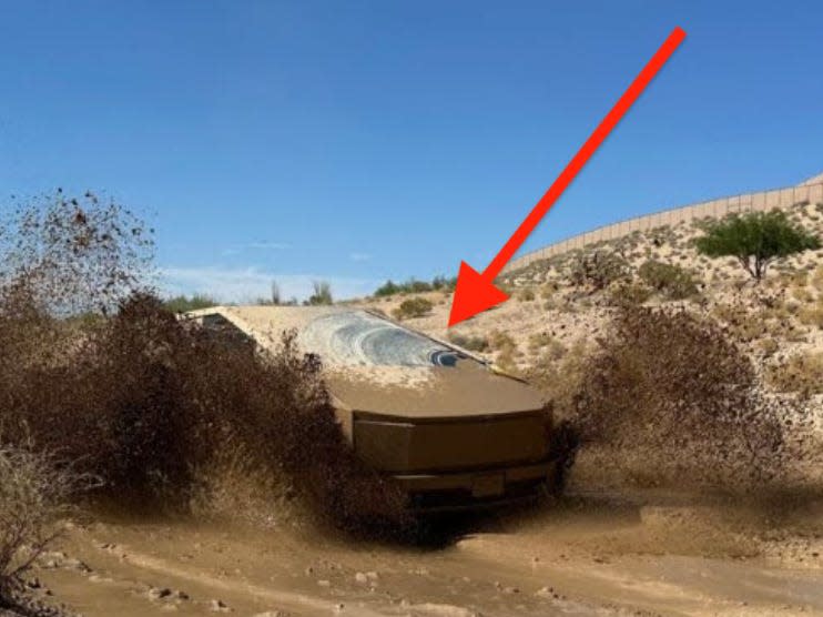 A Cybertruck driving along a dirt track and spraying mud around it with a large red arrow pointing to the windshield wiper where only a section of the window has been cleaned of mud.