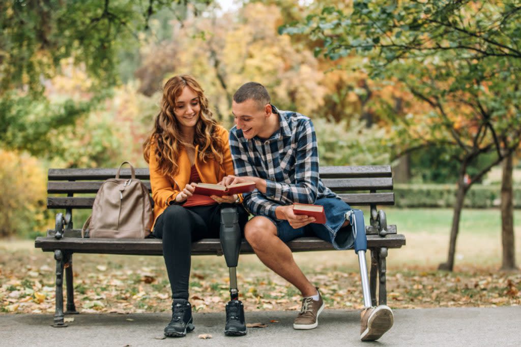 A person with a disability sits on a park bench with a friend as they look at a book with their book bags next to them.