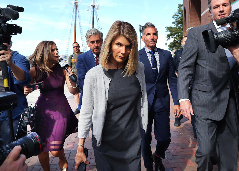 Lori Loughlin walks surrounded by media and security personnel. She wears a grey sweater over a dark dress, holding a purse