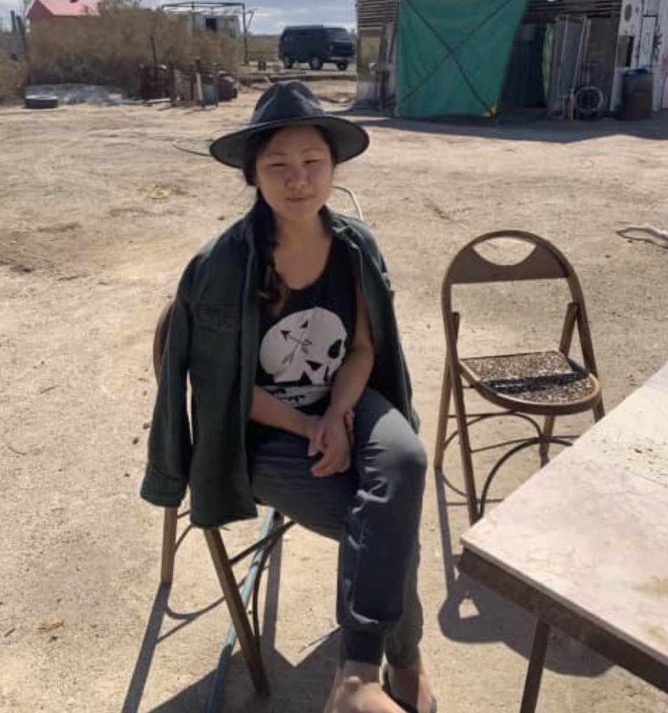 Lauren sitting on a chair wearing light colored pants, a dark tee-shirt with a skull on hit, and a fedora