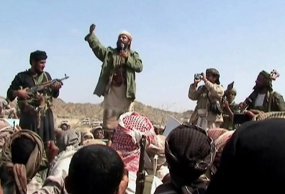 A man holds up a fist while speaking into a microphone to a crowd of people, some of whom wear headdresses and hold guns or cameras.