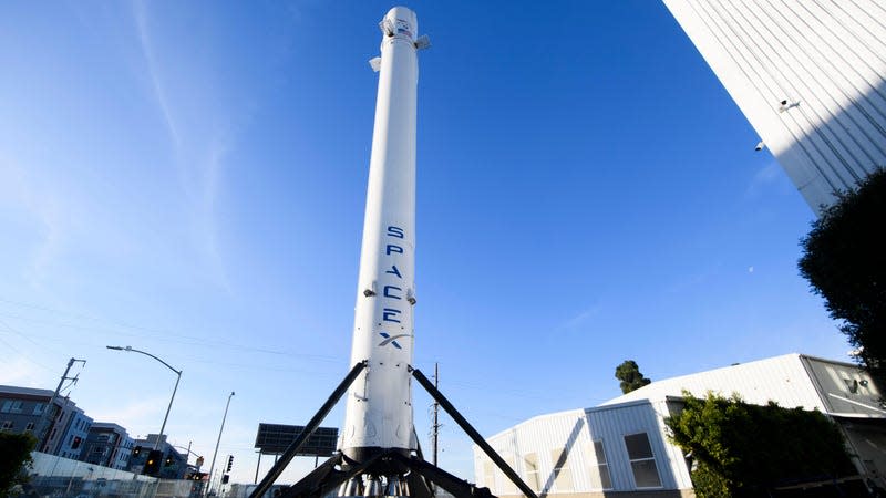 A part of the Falcon 9 Rocket with the SpaceX logo stands up on a large tripod surrounded by nearby buildings